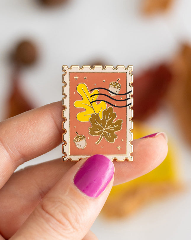 An autumn-themed enamel pin displayed on its branded backing card, styled in a flat-lay with pumpkins, acorns, and fall leaves on a white background.