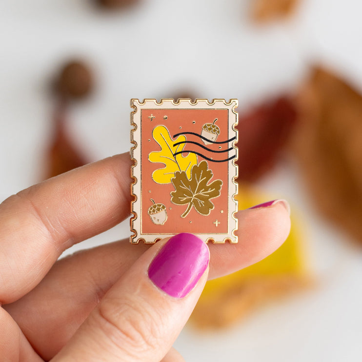 An autumn-themed enamel pin displayed on its branded backing card, styled in a flat-lay with pumpkins, acorns, and fall leaves on a white background.
