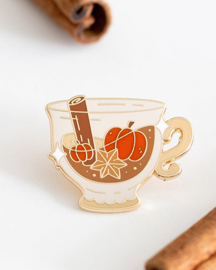 A small enamel pin shaped like a teacup filled with orange pumpkins, cinnamon sticks, and an star anise, displayed on a white surface with cinnamon sticks in the background.