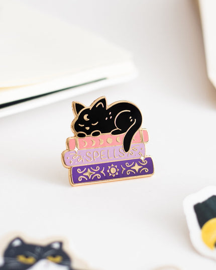 A small enamel pin shaped like a black cat curled up on pink and purple books with the word 'ESPELLER' across the top, displayed on a light surface with other cat-themed items nearby.