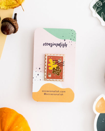 An autumn-themed enamel pin displayed on its branded backing card, styled in a flat-lay with pumpkins, acorns, and fall leaves on a white background.