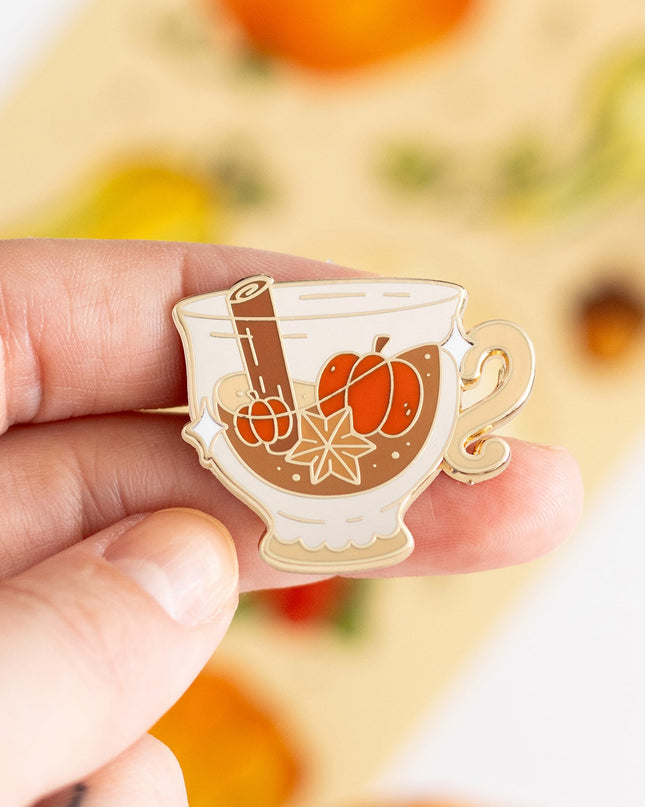 A hand holds a small enamel pin shaped like a teacup filled with orange pumpkin pie filling, cinnamon stick, and star anise, with a gold outline. The background is a soft-focus beige card with autumn illustrations and small pumpkins.