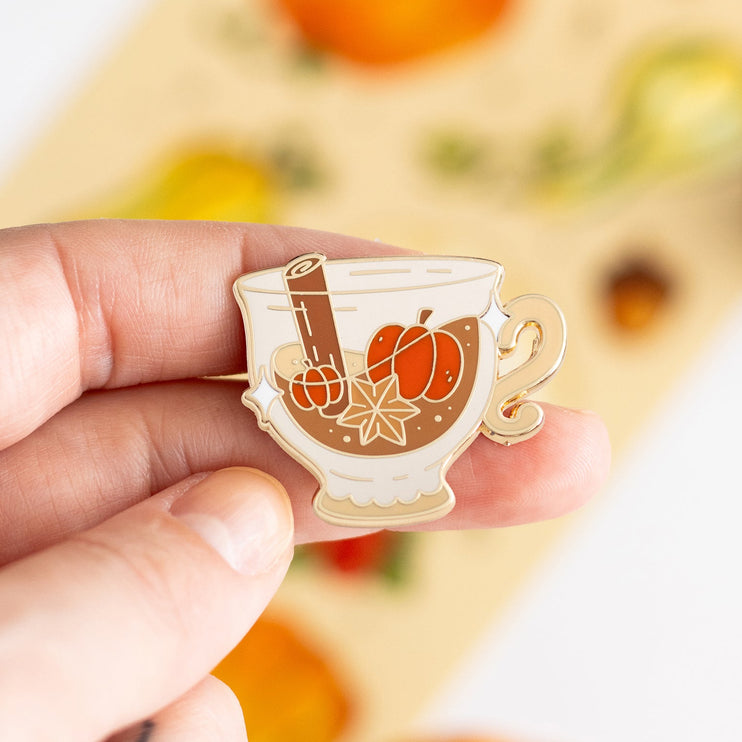 A hand holds a small enamel pin shaped like a teacup filled with orange pumpkin pie filling, cinnamon stick, and star anise, with a gold outline. The background is a soft-focus beige card with autumn illustrations and small pumpkins.