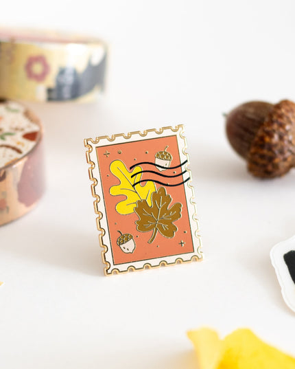 An autumn-themed enamel pin displayed on its branded backing card, styled in a flat-lay with pumpkins, acorns, and fall leaves on a white background.