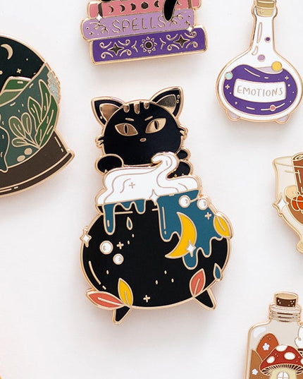 A styled flat-lay of multiple enamel pins with autumn and whimsical designs, including a black cat in a sweater, a potion bottle, a mushroom jar, and a pumpkin illustration, arranged on a white surface with cinnamon sticks, a small pumpkin, acorns, a leaf, and patterned round items in the background.