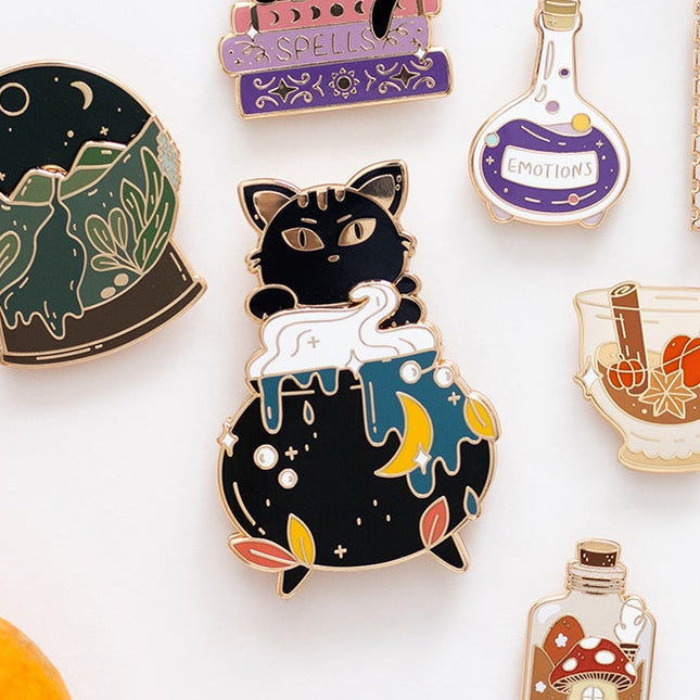 A styled flat-lay of multiple enamel pins with autumn and whimsical designs, including a black cat in a sweater, a potion bottle, a mushroom jar, and a pumpkin illustration, arranged on a white surface with cinnamon sticks, a small pumpkin, acorns, a leaf, and patterned round items in the background.
