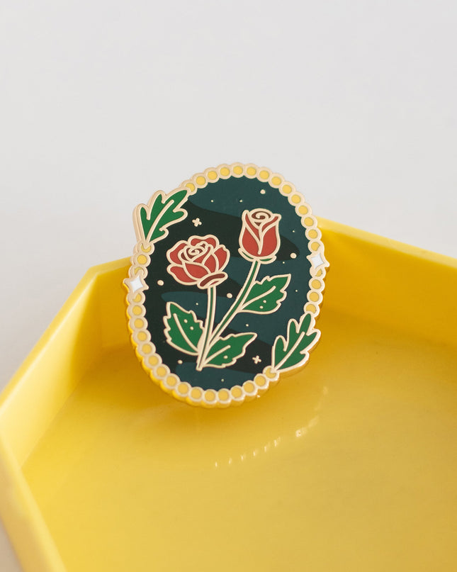 An oval enamel pin featuring two red roses on a dark green background with green leaves and a scalloped gold border, displayed on a yellow hexagonal tray against a light gray background.