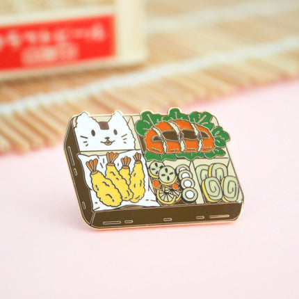 1.5 inch gold plated hard enamel pin of a bento box with pork katsu, tempura shrimp and vegetables, rolled egg omelette and rice decorated as a cat